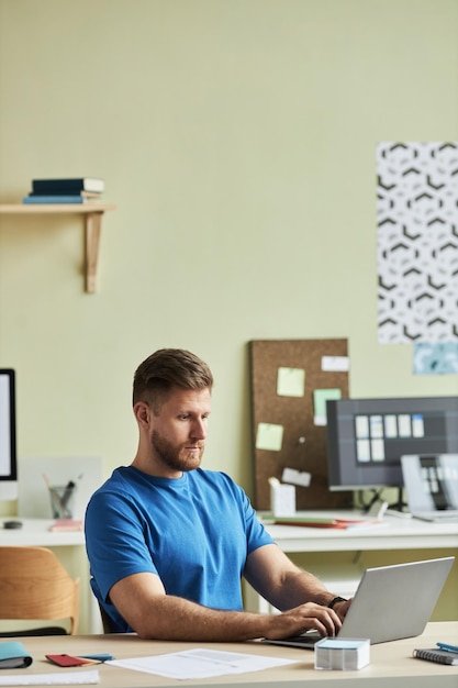Retrato vertical de un joven barbudo que usa una computadora portátil mientras trabaja en un escritorio en un entorno de oficina mínimo