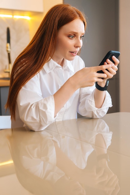 Retrato vertical de una joven pelirroja feliz y atractiva comprobando el contenido del teléfono móvil sentado en