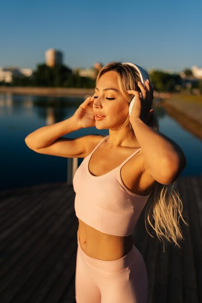 Retrato vertical de una joven rubia con los ojos cerrados en ropa deportiva poniéndose auriculares inalámbricos para empezar a correr en el parque de la ciudad en la mañana de verano