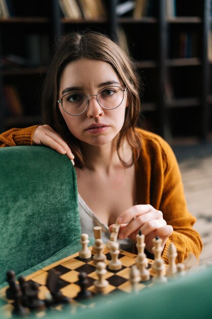 Retrato vertical de una joven seria con anteojos elegantes haciendo que el ajedrez se mueva con una pieza de caballero sentada en el suelo mirando la cámara