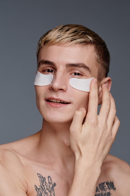 Retrato vertical de un joven tatuado que usa parches faciales hidratantes y sonríe a la cámara mientras disfruta de la rutina masculina de cuidado de la piel