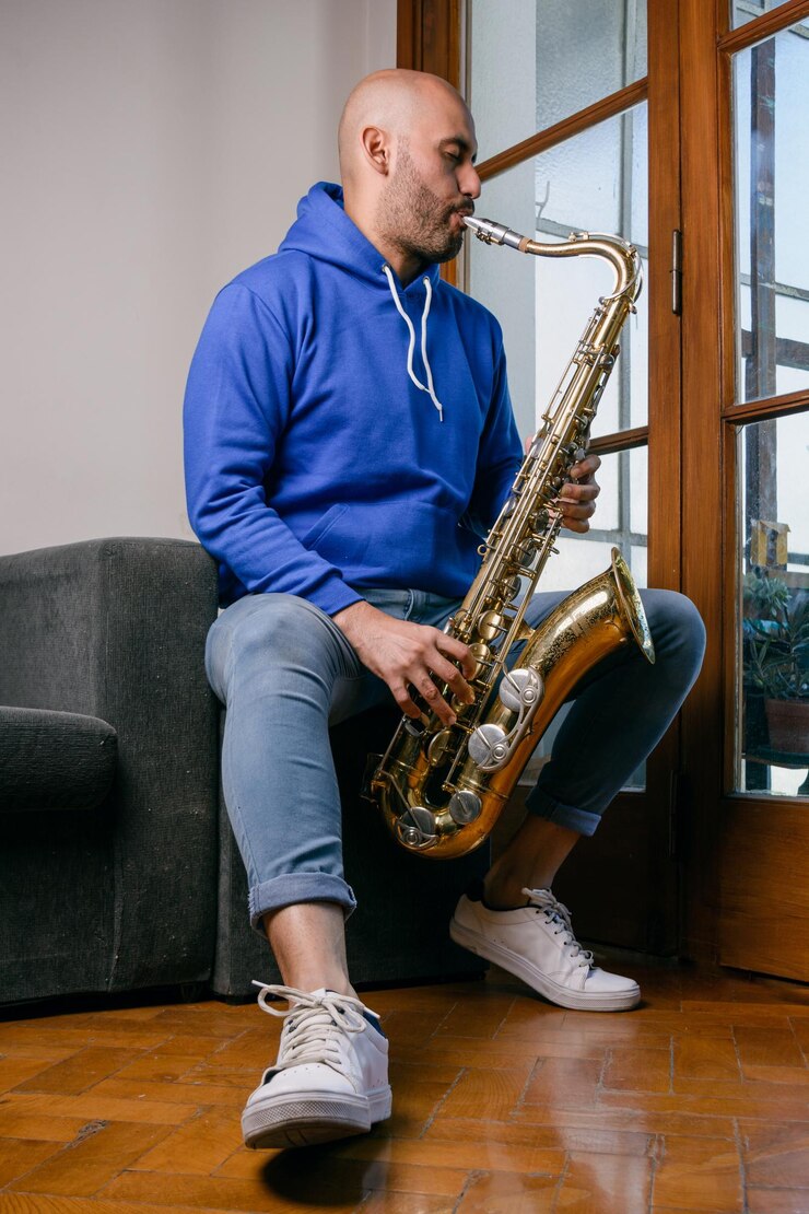 Foto retrato vertical de un joven venezolano vestido de azul tocando el saxofón sentado en el sofá de su casa