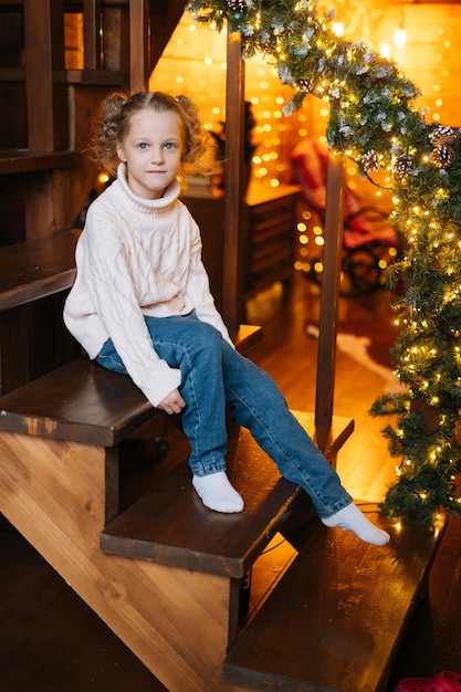 Retrato vertical de una linda niña rubia rizada mirando a la cámara sentada en las escaleras esperando para abrir regalos en la mañana de Navidad