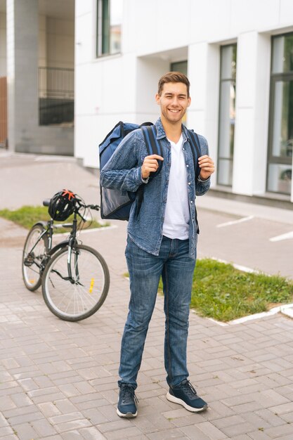 Retrato vertical de longitud completa de alegre apuesto joven repartidor con mochila termo de pie en la calle de la ciudad, en el fondo de la bicicleta y el edificio de oficinas, mirando a la cámara.