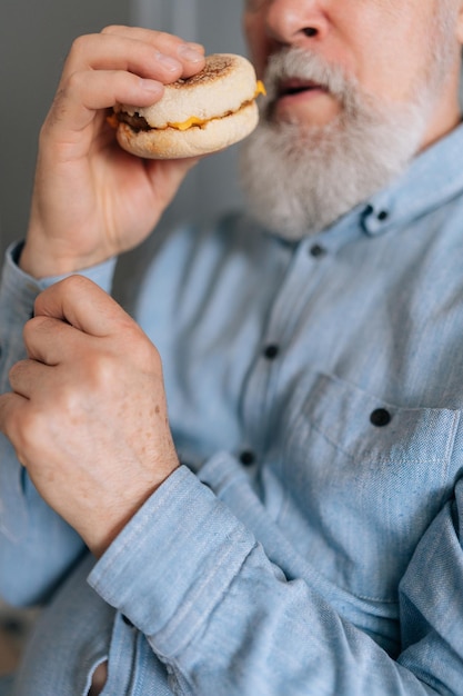 Retrato vertical de un macho adulto maduro descontento comiendo una hamburguesa poco apetecible en la cocina mirando a la cámara con expresión frustrada