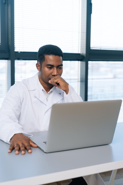 Retrato vertical de un médico practicante negro reflexionando con uniforme blanco pensando mirando la pantalla de la computadora portátil sentado en el escritorio