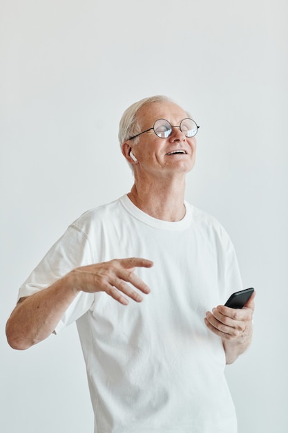 Foto retrato vertical mínimo del hombre mayor sonriente bailando y sosteniendo el teléfono inteligente contra el fondo blanco.