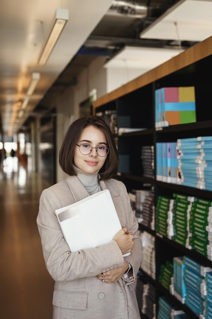 Foto retrato vertical de una mujer guapa inteligente tutora o profesora de pie biblioteca o campus universitario llevar libros para estudiar y aprender material nuevo asistir a cursos de idiomas