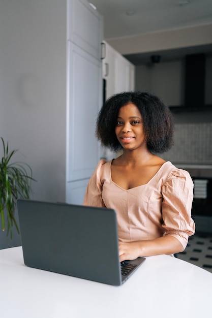 Retrato vertical de una mujer independiente negra positiva sentada a la mesa en la cocina moderna trabajando en una laptop sonriendo mirando la cámara