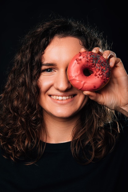 Retrato vertical de una mujer rizada que cubre un ojo con un donut rosa