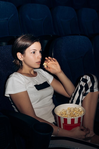 Retrato vertical de una mujer sentada en una silla en un teatro de cine comiendo un cuenco de palomitas de maíz