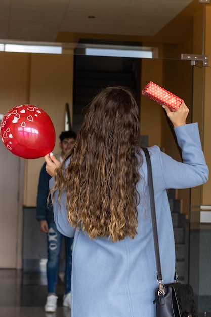 Retrato vertical de niña esperando a su novio en la puerta de su casa para sorprenderlo con un regalo y un globo para celebrar un día especial. San Valentín, aniversario, cumpleaños...