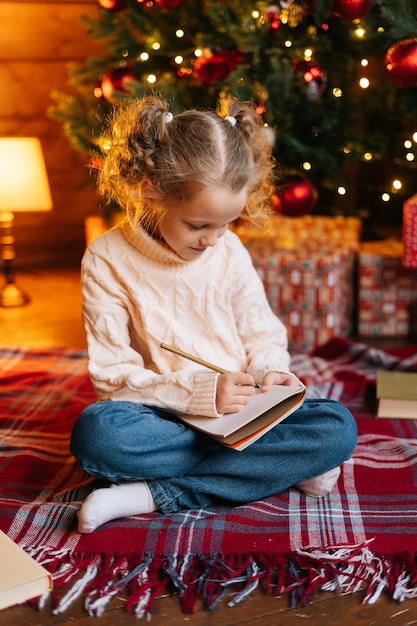 Retrato vertical de una pequeña niña rubia y rizada enfocada escribiendo una carta a Santa Claus sentada en el suelo sobre el fondo del árbol de Navidad y cajas de regalo