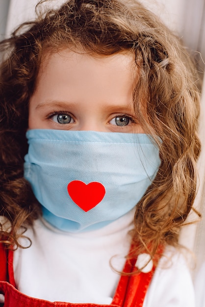 Retrato vertical recortado de una niña linda europea con una máscara de salud con un corazón rojo como una forma de mostrar agradecimiento y agradecer a todos los empleados esenciales durante la pandemia de Covid-19.