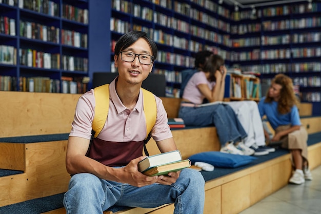 Foto retrato vibrante de un joven asiático sosteniendo libros en la biblioteca de la universidad y sonriendo al espacio de copia de la cámara