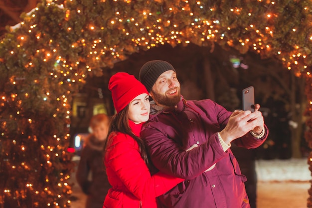 Retrato de vista cercana de pareja joven haciendo selfie cerca del arco de abeto, decorado con guirnaldas. Pareja casada fotografiando mientras viaja de vacaciones de invierno