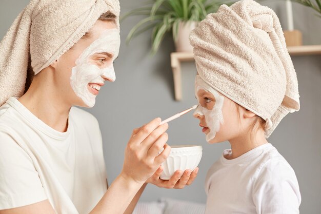 Retrato de vista lateral de una familia alegre haciendo procedimientos de cosmetología disfrutando de pasar tiempo juntos en casa mujer aplicando máscara a su hija