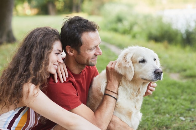 Retrato de vista lateral de la feliz pareja joven con perro sentado sobre la hierba verde al aire libre