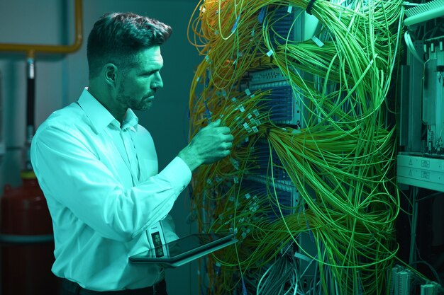 Retrato de vista lateral del guapo ingeniero de datos inspeccionando cables en la sala de servidores mientras trabaja con la supercomputadora en luz azul, espacio de copia