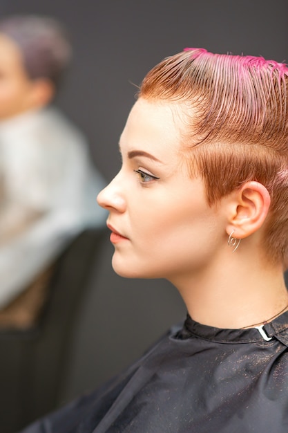 Retrato de vista lateral de una hermosa joven caucásica con un corte de pelo corto de color rosa esperando una peluquería en un salón de belleza