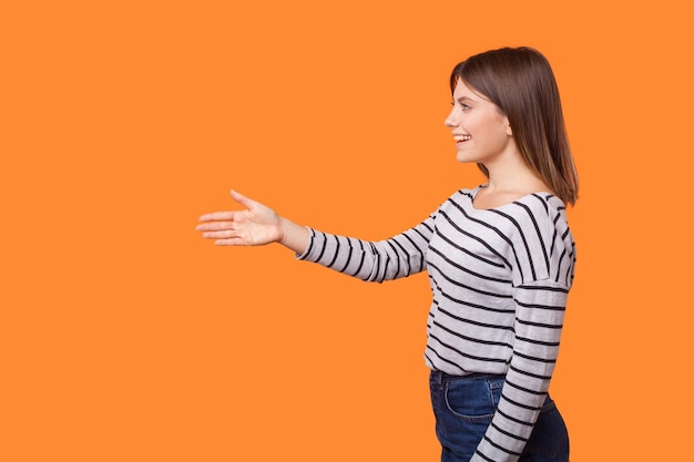 Foto retrato de vista lateral de una hermosa mujer amigable con cabello castaño en camisa a rayas de manga larga de pie, dando la mano para el apretón de manos, saludando con una sonrisa. tiro de estudio interior aislado sobre fondo naranja