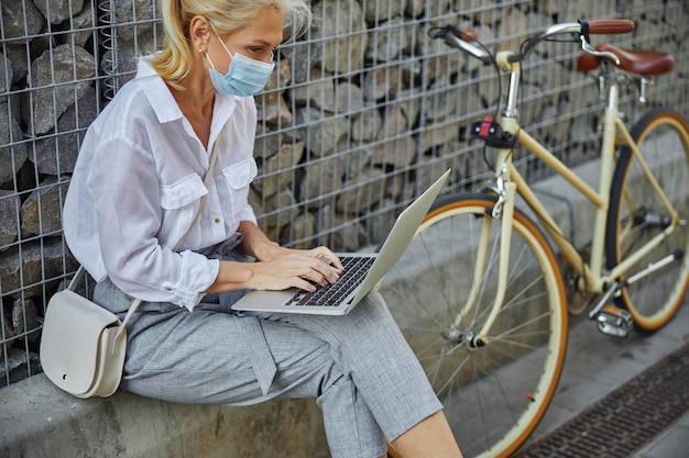 Retrato de vista lateral de hermosa mujer de negocios inteligente en máscara protectora trabajando en la computadora portátil mientras bicicleta retro de pie cerca de ella al aire libre