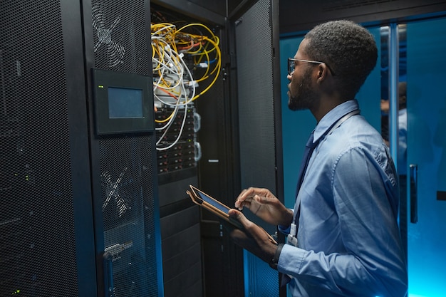 Retrato de vista lateral del hombre afroamericano de pie junto al gabinete del servidor mientras trabaja con la supercomputadora en el centro de datos y sosteniendo la tableta, espacio de copia