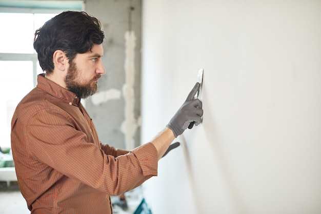 Retrato de vista lateral del hombre barbudo alisando la pared blanca con una espátula mientras se renueva la casa, espacio de copia