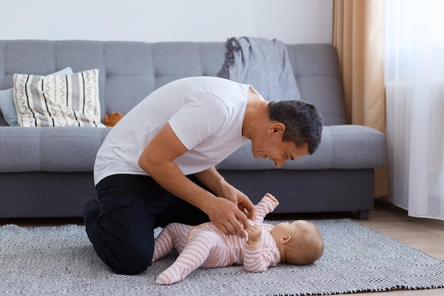 Retrato de vista lateral de hombre feliz con cabello oscuro con camiseta blanca jugando con su hija pequeña mientras está sentado en el piso cerca del sofá y la ventana en la sala de estar luminosa.