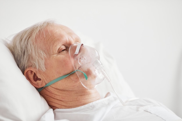 Retrato de vista lateral del hombre mayor enfermo acostado en la cama de un hospital con máscara de suplemento de oxígeno, espacio de copia