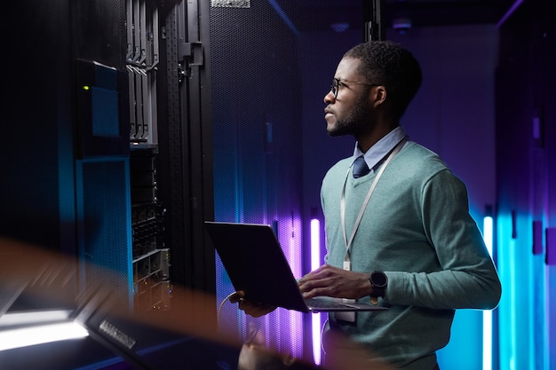 Retrato de vista lateral del ingeniero de datos afroamericano sosteniendo la computadora portátil mientras trabaja con la supercomputadora en la sala de servidores iluminada por la luz azul, espacio de copia