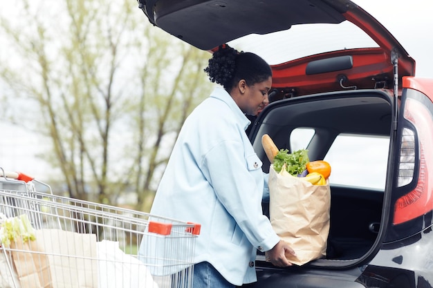 Retrato de vista lateral de una joven negra poniendo una bolsa de supermercado en el maletero del auto en el estacionamiento del supermercado