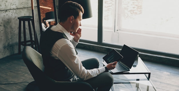Retrato de vista lateral de un joven vestido con traje de propietario sentado en su oficina trabajando mientras habla en el teléfono inteligente y mirando a una pantalla de tableta.