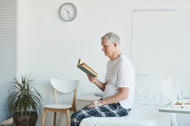 Retrato de vista lateral del libro de lectura del hombre mayor en el hospital mientras está sentado en la cama en el espacio de copia de la habitación blanca