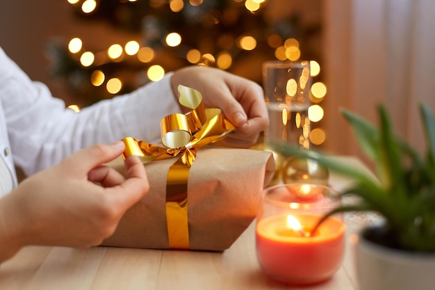 Retrato de vista lateral de manos femeninas anónimas desconocidas sosteniendo una caja de regalo envuelta con un árbol de Navidad decorado con luces en la caja de regalo de embalaje de fondo con lazo dorado