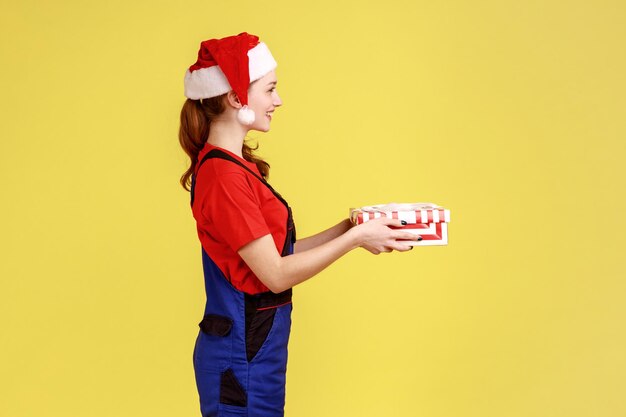 Retrato de vista lateral de una mensajera sonriente de pie dando una caja de regalo envuelta para el cliente, usando un mono azul y un sombrero de santa claus. Disparo de estudio interior aislado sobre fondo amarillo.