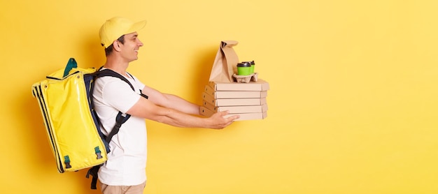 Retrato de vista lateral de un mensajero feliz con camiseta y gorra sosteniendo cajas de pizza y bebida para llevar posando aislado sobre fondo amarillo entregando pedidos puerta a puerta