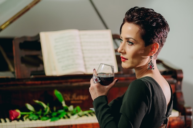 Retrato de vista lateral de una mujer atractiva y elegante sentada al piano y sosteniendo una copa de vino Rosa roja en el teclado del piano