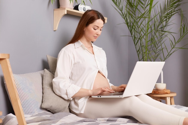 Retrato de vista lateral de una mujer caucásica de cabello castaño embarazada sentada en la cama y trabajando en una computadora portátil escribiendo en el teclado respondiendo correos electrónicos comunicándose en línea en las redes sociales