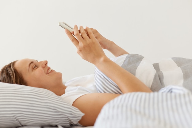 Retrato de vista lateral de mujer feliz satisfecha con camiseta blanca casual acostada debajo de la manta en la cama, usando el teléfono inteligente, mirando la pantalla del teléfono móvil con una sonrisa positiva y agradable.