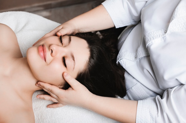 Retrato de vista lateral de una mujer increíble apoyado en una cama de spa con los ojos cerrados con terapia de cuidado de la piel facial por una cosmetóloga en un centro de spa