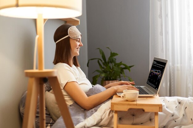 Retrato de vista lateral de una mujer independiente feliz sonriente que usa máscara para dormir sentada con una computadora portátil en la cama en casa y escribiendo en el teclado trabajando en línea por la mañana