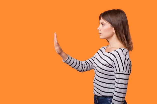 Retrato de vista lateral de una mujer joven preocupada con cabello castaño en camisa a rayas de manga larga de pie levantando el brazo mostrando el gesto de parada señal de advertencia estudio interior tiro aislado sobre fondo naranja