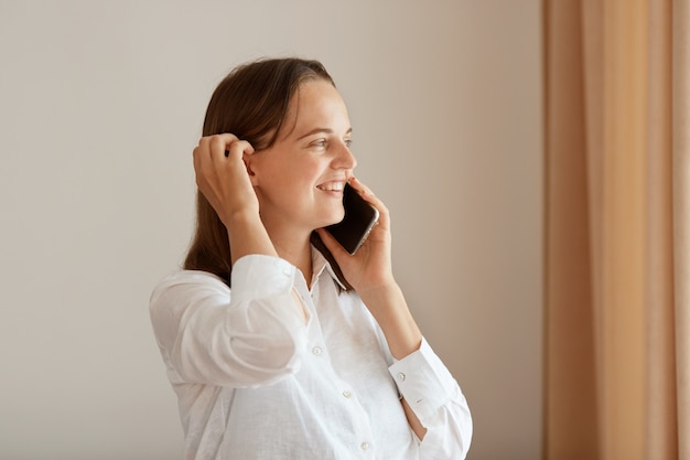 Retrato de vista lateral de mujer positiva de pie junto a la ventana con cortinas beige, joven adulta con camisa de algodón blanca hablando por teléfono celular, escuchando buenas noticias.