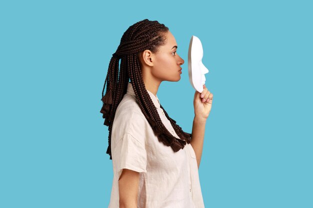 Retrato de vista lateral de una mujer con rastas negras sosteniendo una máscara blanca frente a su rostro con una expresión facial estricta usando una camisa blanca Disparo de estudio interior aislado sobre fondo azul