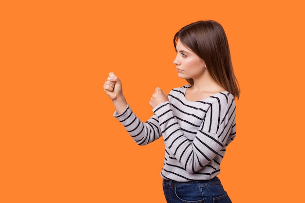 Retrato de vista lateral de una mujer seria y segura de sí misma con cabello castaño en camisa a rayas de manga larga apretando los puños mostrando un gesto de boxeo y lista para perforar una foto de estudio interior aislada en un fondo naranja