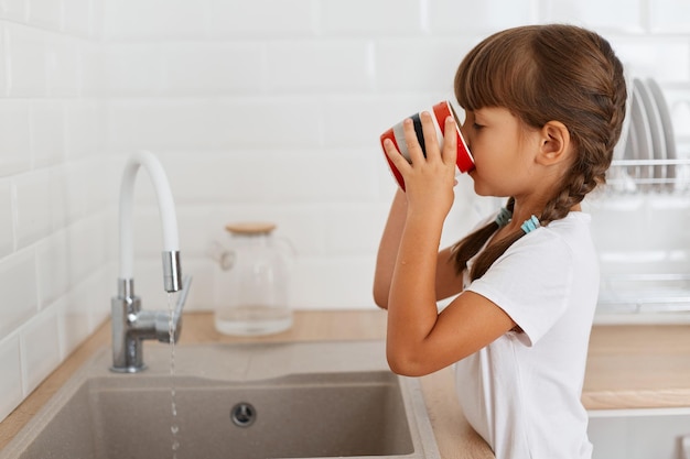 Retrato de vista lateral de una niña de cabello oscuro con coletas usando una camiseta blanca sosteniendo una taza y de pie cerca del grifo de la cocina bebiendo agua limpia siente sed