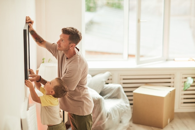 Retrato de vista lateral de padre e hijo colgando cuadros en la pared mientras se muda al nuevo espacio de copia de casa