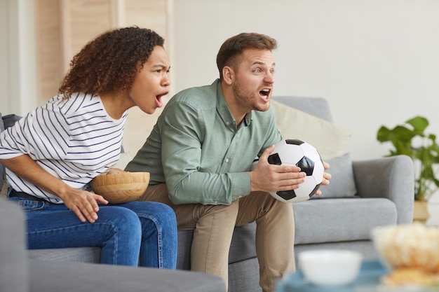 Retrato de vista lateral de una pareja emocional viendo partidos deportivos en la televisión en casa y animando en voz alta mientras está sentado en el sofá en el acogedor apartamento