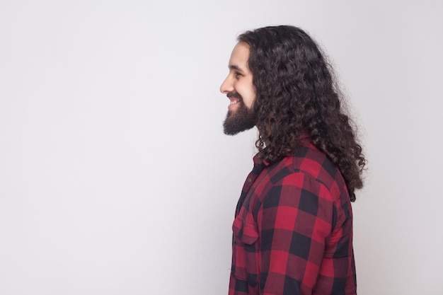 Retrato de vista lateral de perfil de hombre feliz guapo con barba y pelo largo y rizado negro en estilo casual, camisa roja a cuadros de pie con una gran sonrisa. tiro del estudio de interior, aislado en fondo gris.